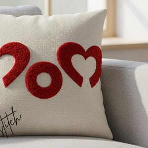 Op de afbeelding: Crèmekleurig decoratief kussen met het woord "MOM" in rode, hartvormige letters. Het kussen ligt op een grijze bank. Het woord "Jostitch" is cursief geschreven in de linkerbenedenhoek van het kussen.
