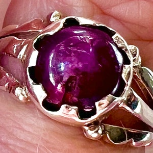 Op de afbeelding: Een zilveren ring met een grote, gefacetteerde, paarse edelsteen gezet in een gedetailleerde, sierlijke zetting.