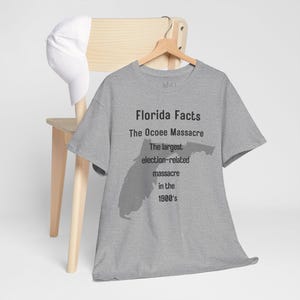 Può includere: Una maglietta grigio melange con la scritta "Florida Facts" e informazioni sul massacro di Ocoee. La maglietta presenta una silhouette dello stato della Florida. Un berretto da baseball bianco è appoggiato su una sedia di legno.
