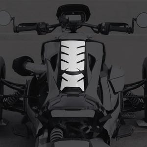 K&ouml;nnte beinhalten: Ein wei&szlig;er Aufkleber mit einem segmentierten Design ist an der Vorderseite eines dreir&auml;drigen Motorrads angebracht. Das Motorrad hat schwarze Lenker, Spiegel und Reifen. Das Bild ist in Graustufen.