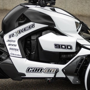 K&ouml;nnte beinhalten: Ein wei&szlig;-schwarzes Can-Am Ryker 900 Dreirad-Motorrad mit schwarzen und wei&szlig;en Grafiken. Das Motorrad hat einen schwarzen Sitz und einen schwarzen Auspuff.