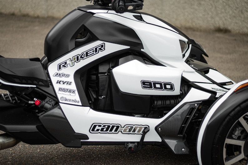 K&ouml;nnte beinhalten: Ein wei&szlig;-schwarzes Can-Am Ryker 900 Dreirad-Motorrad mit schwarzen und wei&szlig;en Grafiken. Das Motorrad hat einen schwarzen Sitz und einen schwarzen Auspuff.