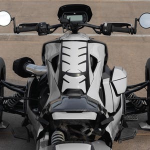 K&ouml;nnte beinhalten: Ein wei&szlig;-schwarzes dreir&auml;driges Motorrad mit einem einzigartigen Design. Das Fahrzeug hat einen schwarzen Lenker, Seitenspiegel und ein schwarz-wei&szlig;es Muster auf der Vorderseite. Die Reifen sind wei&szlig; mit schwarzen Akzenten. Das Motorrad steht auf einer Betonoberfl&auml;che.