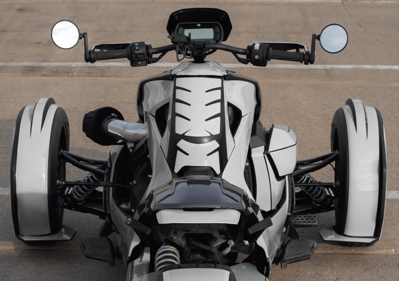 K&ouml;nnte beinhalten: Ein wei&szlig;-schwarzes dreir&auml;driges Motorrad mit einem einzigartigen Design. Das Fahrzeug hat einen schwarzen Lenker, Seitenspiegel und ein schwarz-wei&szlig;es Muster auf der Vorderseite. Die Reifen sind wei&szlig; mit schwarzen Akzenten. Das Motorrad steht auf einer Betonoberfl&auml;che.