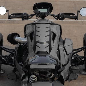 K&ouml;nnte beinhalten: Ein graues Dreirad-Motorrad mit schwarzen Akzenten, das ein schlankes Design aufweist. Das Fahrzeug hat zwei Seitenspiegel, ein digitales Display und eine niedrige Bauweise. Die Reifen haben ein einzigartiges Profil.