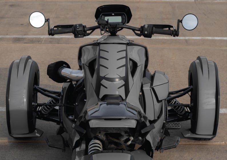 K&ouml;nnte beinhalten: Ein graues Dreirad-Motorrad mit schwarzen Akzenten, das ein schlankes Design aufweist. Das Fahrzeug hat zwei Seitenspiegel, ein digitales Display und eine niedrige Bauweise. Die Reifen haben ein einzigartiges Profil.