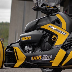 K&ouml;nnte beinhalten: Ein schwarzes und gelbes Can-Am Ryker 900 Dreirad-Motorrad mit einem schwarz-wei&szlig;en "Can-Am"-Logo und dem Text "Ryker" an der Seite.