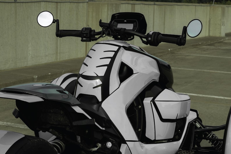 K&ouml;nnte beinhalten: Ein wei&szlig;-schwarzes Dreirad-Motorrad mit modernem Design. Das Fahrzeug hat eine schlanke, kantige Karosserie mit schwarzen Akzenten und runden Seitenspiegeln. Der Lenker und das Armaturenbrett sind sichtbar.