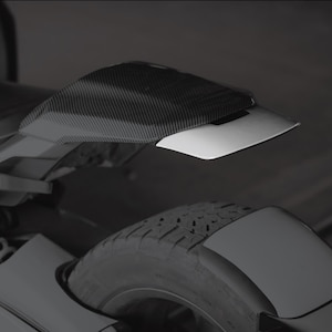 May include: A motorcycle rear fender with a carbon fiber finish and a white accent. The fender is mounted above the rear tire and suspension, providing protection and a sleek aesthetic. The image is in grayscale.