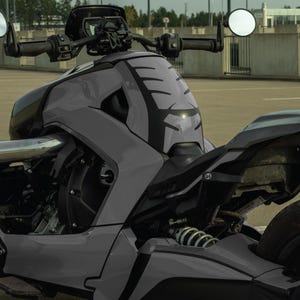 K&ouml;nnte beinhalten: Ein graues dreir&auml;driges Motorrad mit schwarzen Akzenten. Das Fahrzeug hat ein schlankes, modernes Design mit niedrigem Profil, mit einer markanten Frontverkleidung, runden Spiegeln und einem einzelnen Hinterrad. Das Motorrad steht auf einer gepflasterten Fl&auml;che.