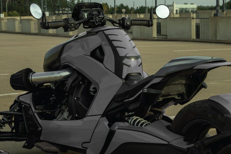 K&ouml;nnte beinhalten: Ein graues dreir&auml;driges Motorrad mit schwarzen Akzenten. Das Fahrzeug hat ein schlankes, modernes Design mit niedrigem Profil, mit einer markanten Frontverkleidung, runden Spiegeln und einem einzelnen Hinterrad. Das Motorrad steht auf einer gepflasterten Fl&auml;che.