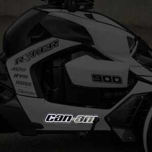 K&ouml;nnte beinhalten: Ein wei&szlig;-schwarzes Can-Am Ryker 900 Dreirad-Motorrad mit reflektierenden Aufklebern. Die Aufkleber enthalten die W&ouml;rter "Ryker", "Can-Am", "900", "XPS", "KYB", "ROTAX" und "BRP".