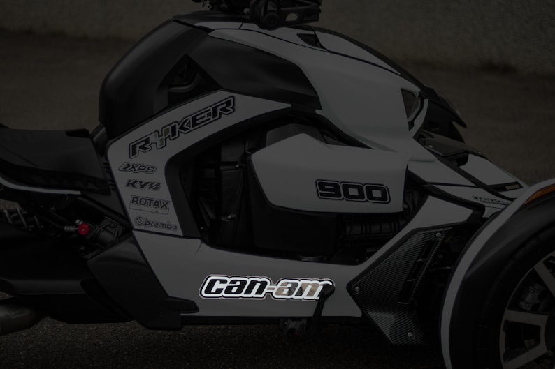 K&ouml;nnte beinhalten: Ein wei&szlig;-schwarzes Can-Am Ryker 900 Dreirad-Motorrad mit reflektierenden Aufklebern. Die Aufkleber enthalten die W&ouml;rter "Ryker", "Can-Am", "900", "XPS", "KYB", "ROTAX" und "BRP".