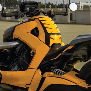 K&ouml;nnte beinhalten: Ein gelb-schwarzes Dreirad-Motorrad mit einzigartigem Design. Das Fahrzeug verf&uuml;gt &uuml;ber einen schwarzen Sitz, einen Lenker mit runden Spiegeln und eine markante Frontverkleidung. Das Motorrad steht auf einer gepflasterten Fl&auml;che.