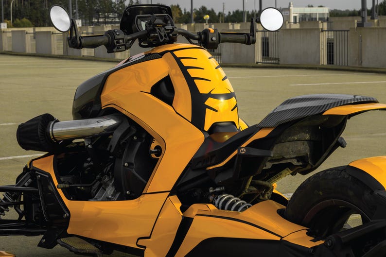 K&ouml;nnte beinhalten: Ein gelb-schwarzes Dreirad-Motorrad mit einzigartigem Design. Das Fahrzeug verf&uuml;gt &uuml;ber einen schwarzen Sitz, einen Lenker mit runden Spiegeln und eine markante Frontverkleidung. Das Motorrad steht auf einer gepflasterten Fl&auml;che.