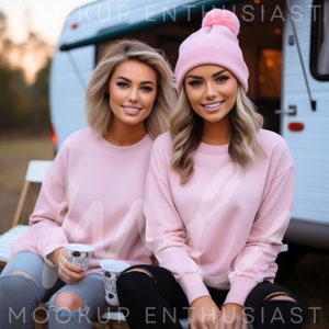 May include: Two women wearing matching pink sweatshirts and a pink beanie with a pom-pom. One woman is holding a white cup with black text. The other woman is sitting next to her. They are sitting on a white bench in front of a camper.