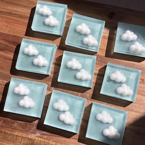 Puede incluir: Ocho pastillas de jabón cuadradas de color azul claro translúcido, cada una con dos formas de nube blancas tridimensionales incrustadas. Las pastillas están dispuestas en un patrón de cuadrícula, mostrando un acabado liso semitransparente.