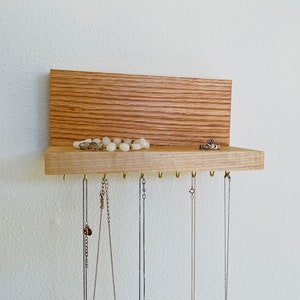 Pu&ograve; includere: Un organizzatore per gioielli in legno con uno scaffale e ganci per appendere collane. L'organizzatore &egrave; realizzato in legno marrone chiaro e ha una finitura in legno naturale. Ci sono diversi collane appese ai ganci, e un anello e una collana sono sullo scaffale.