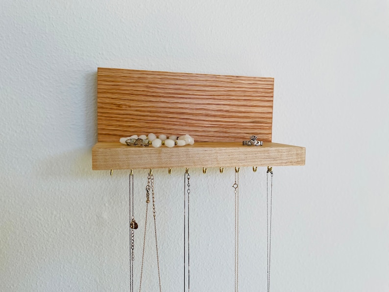 Pu&ograve; includere: Un organizzatore per gioielli in legno con uno scaffale e ganci per appendere collane. L'organizzatore &egrave; realizzato in legno marrone chiaro e ha una finitura in legno naturale. Ci sono diversi collane appese ai ganci, e un anello e una collana sono sullo scaffale.