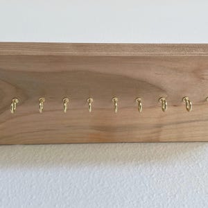 Pu&ograve; includere: Un portachiavi in legno con 10 ganci dorati. Il portachiavi &egrave; in legno marrone chiaro e ha una forma rettangolare.
