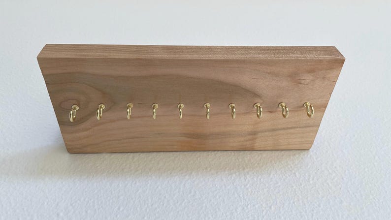 Pu&ograve; includere: Un portachiavi in legno con 10 ganci dorati. Il portachiavi &egrave; in legno marrone chiaro e ha una forma rettangolare.