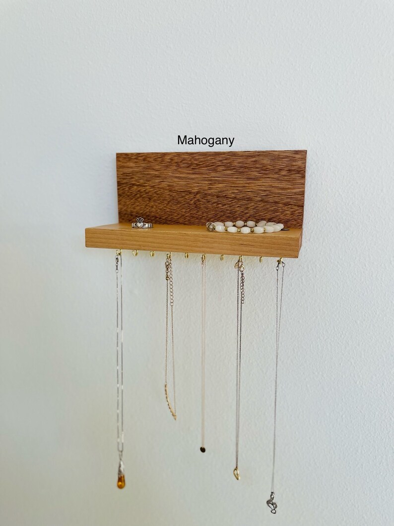 Pu&ograve; includere: Un organizzatore per gioielli in legno di mogano con uno scaffale e ganci per appendere collane. L'organizzatore ha un'etichetta che dice "Mogano".