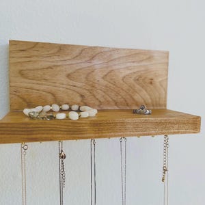 Pu&ograve; includere: Un organizzatore per gioielli in legno con uno scaffale e ganci per appendere collane e braccialetti. Una collana di perle bianche e un anello d'argento sono esposti sullo scaffale.