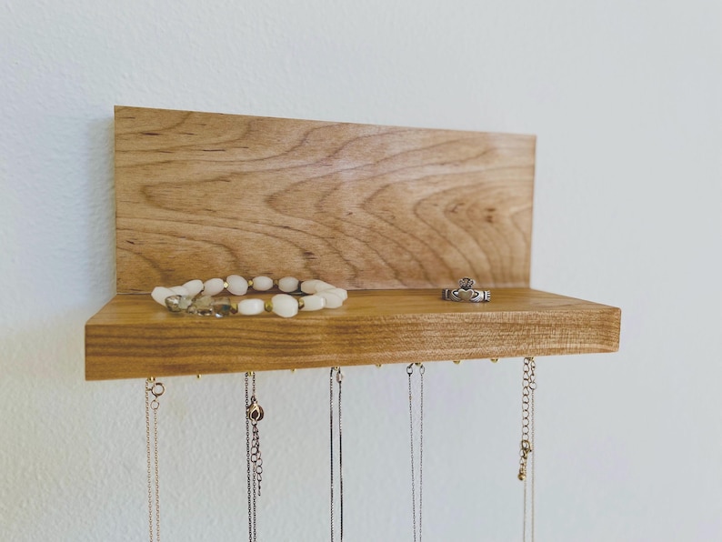 Pu&ograve; includere: Un organizzatore per gioielli in legno con uno scaffale e ganci per appendere collane e braccialetti. Una collana di perle bianche e un anello d'argento sono esposti sullo scaffale.