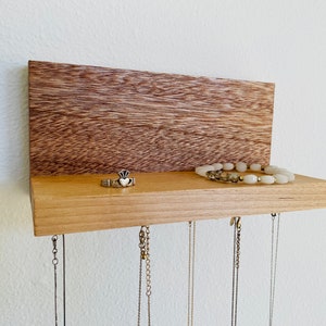 Pu&ograve; includere: Un organizzatore per gioielli in legno con uno scaffale e ganci. Lo scaffale &egrave; in legno chiaro e il retro &egrave; in legno scuro. C'&egrave; un anello e un braccialetto sullo scaffale. Ci sono diverse collane appese ai ganci.