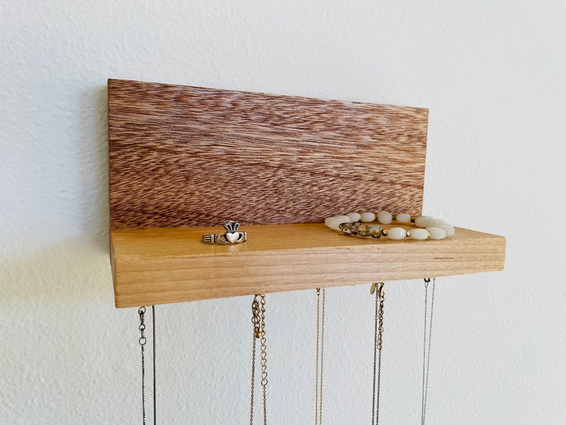 Pu&ograve; includere: Un organizzatore per gioielli in legno con uno scaffale e ganci. Lo scaffale &egrave; in legno chiaro e il retro &egrave; in legno scuro. C'&egrave; un anello e un braccialetto sullo scaffale. Ci sono diverse collane appese ai ganci.