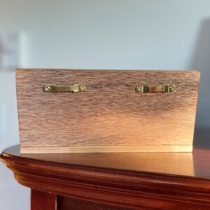 Pu&ograve; includere: Il retro di una scatola rettangolare in legno con due pezzi di ferramenta dorati per l'appeso fissati nella parte superiore.