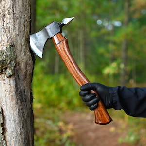 Könnte beinhalten: Eine Person mit schwarzen Handschuhen hält eine handgeschmiedete Axt mit einem Holzstiel. Die Axt ist teilweise in einen Baumstamm geschlagen.