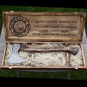 May include: A wooden box with a personalized axe inside, designed for a firefighter. The box lid is engraved with the words "ALEX CARTER - BADGE 2741" and a fire department emblem. The axe has the words "BRAVERY IS YOUR EVERYDAY" engraved on it.