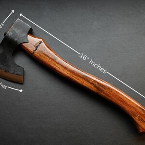 May include: A handcrafted axe with a dark metal head and a wooden handle. The axe head is 5.5 inches wide, and the handle is 16 inches long. The axe has a curved handle and a dark finish, suitable for outdoor activities.
