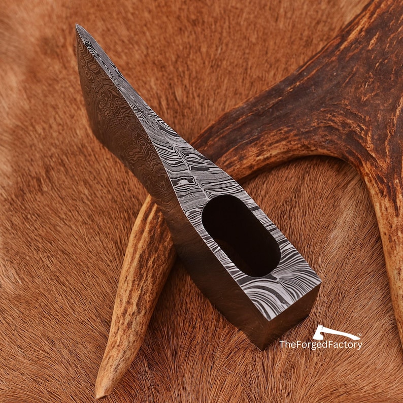 K&ouml;nnte beinhalten: Ein handgeschmiedeter Axtkopf mit einem Damastmesser-Klingenmuster. Der Axtkopf liegt auf einer braunen Fellfl&auml;che mit einem St&uuml;ck Geweih.