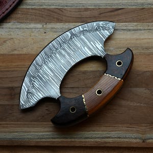 May include: A crescent-shaped ulu knife featuring a patterned Damascus steel blade and a multi-toned wooden handle. The handle is constructed from two distinct types of wood, secured with three brass rivets, and features a curved grip design.