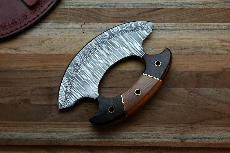 May include: A crescent-shaped ulu knife featuring a patterned Damascus steel blade and a multi-toned wooden handle. The handle is constructed from two distinct types of wood, secured with three brass rivets, and features a curved grip design.