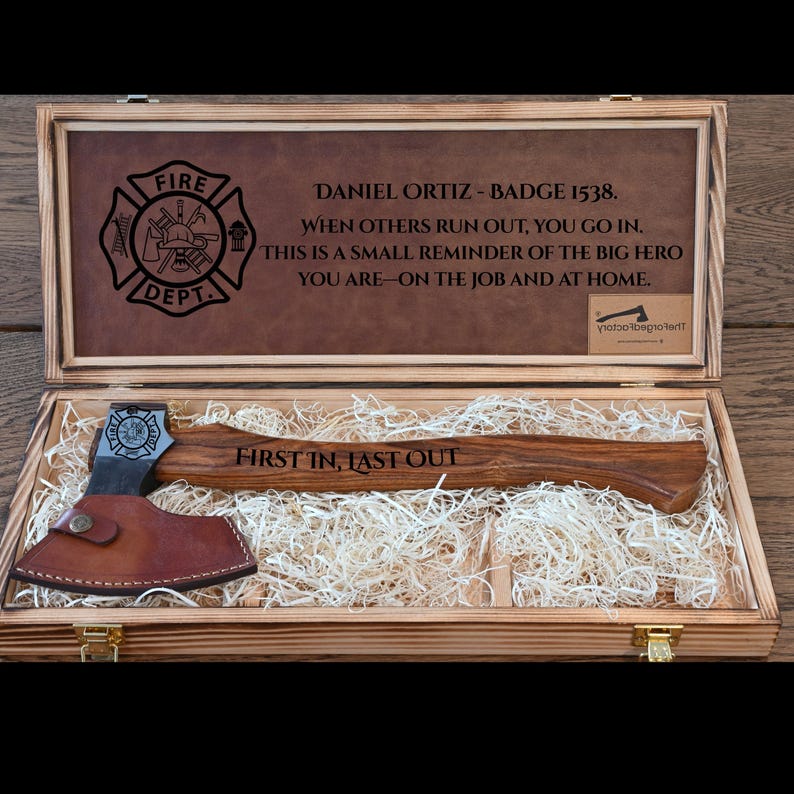 May include: A wooden presentation box with a firefighter's axe. The axe has a wooden handle and a metal head with a leather sheath. The box contains text and a firefighter's emblem. The text on the axe reads "FIRST IN, LAST OUT."