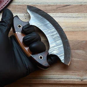 May include: A crescent-shaped ulu knife featuring a patterned Damascus steel blade and a dark brown wooden handle with two brass rivets. The handle is constructed from two distinct wood tones, showcasing a smooth finish and a curved grip design.