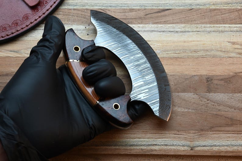 May include: A crescent-shaped ulu knife featuring a patterned Damascus steel blade and a dark brown wooden handle with two brass rivets. The handle is constructed from two distinct wood tones, showcasing a smooth finish and a curved grip design.