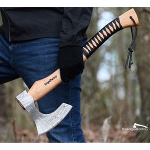 May include: A hand axe with a patterned Damascus steel blade and a light-colored wooden handle. The handle features a black leather wrap in a crisscross pattern, a black leather lanyard at the end, and the text ForgedFactory printed on the wood.