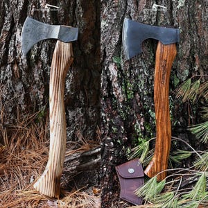 May include: Two axes with wooden handles and metal blades are displayed against a tree trunk. One axe has a patterned blade, the other has a solid blade. A leather sheath is visible near the second axe. The handles have a curved design.