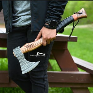 Puede incluir: Un hacha artesanal con mango de madera y hoja de acero estampada. El mango está envuelto en cuero negro y asegurado con una correa de cuero. El hacha se sostiene contra un pantalón y una chaqueta negros. El texto "TheForgedFactory" es visible.