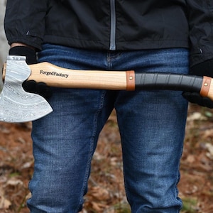 May include: Axe with a wooden handle and a patterned steel blade. The handle is wrapped in black leather with brown leather accents. The text "ForgedFactory" is printed on the wooden handle.