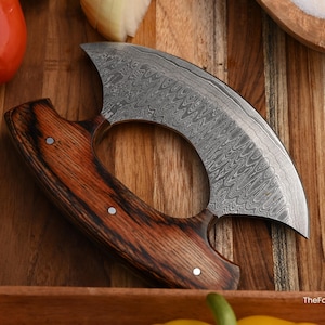 May include: A crescent-shaped ulu knife featuring a Damascus steel blade with a distinct wavy pattern and a dark brown wood handle secured with three metal rivets. The text TheForgedFactory is visible in the bottom right corner.