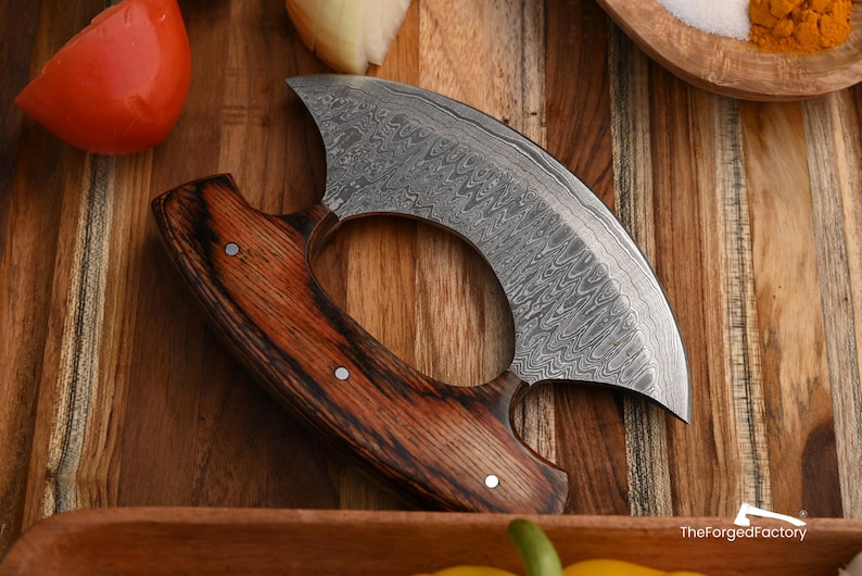 May include: A crescent-shaped ulu knife featuring a Damascus steel blade with a distinct wavy pattern and a dark brown wood handle secured with three metal rivets. The text TheForgedFactory is visible in the bottom right corner.