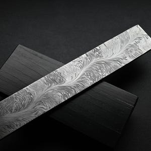 May include: A rectangular, patterned metal blade rests on two dark wooden blocks. The blade features a swirling, feather-like design in shades of silver and gray. The blocks are rectangular and have a dark, matte finish.