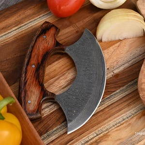 May include: A crescent-shaped mezzaluna knife with a dark, patterned Damascus steel blade and a polished wooden handle secured with three metal rivets. The blade features a distinct wavy texture, and the text TheForgedFactory is visible.