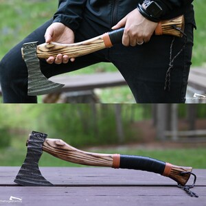 May include: A hand axe with a dark, textured, forged metal head featuring a wavy pattern. The handle is crafted from light-colored wood with prominent dark grain, accented by a black leather grip, tan leather bands, and a black cord lanyard.