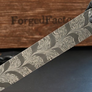 May include: A rectangular metal blade with a decorative floral pattern in shades of grey and white is held by a gloved hand. The blade is in front of a wooden box with the text "Forged Factory" on it. The blade is likely for a knife.